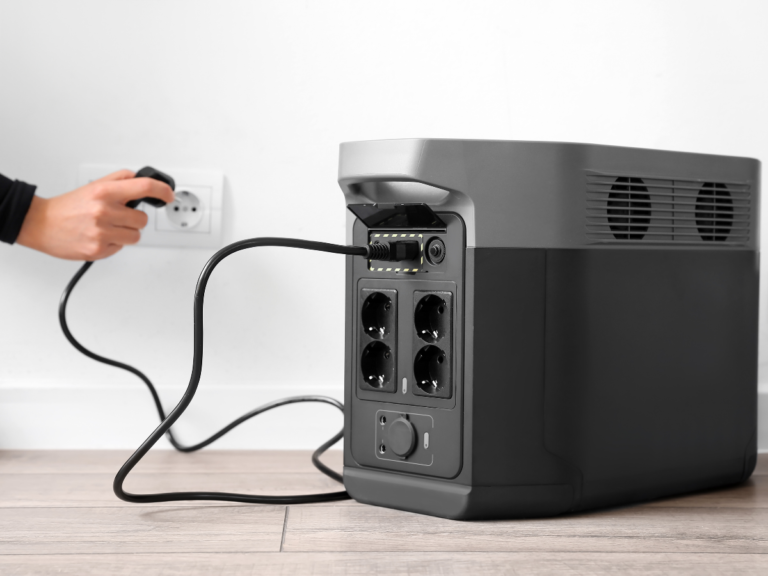 Hand plugging a black cable into a wall socket, connected to a large portable battery unit standing on a wooden floor