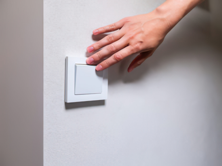 Hand reaching towards a white wall light switch on a pale grey wall