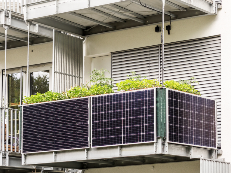 Three solar panels mounted along the railing of an apartment balcony with green plants growing behind them