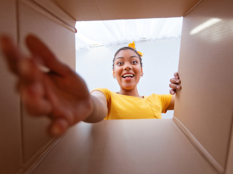 Smiling woman in a yellow t-shirt reaching into an open cardboard box, seen from inside the box